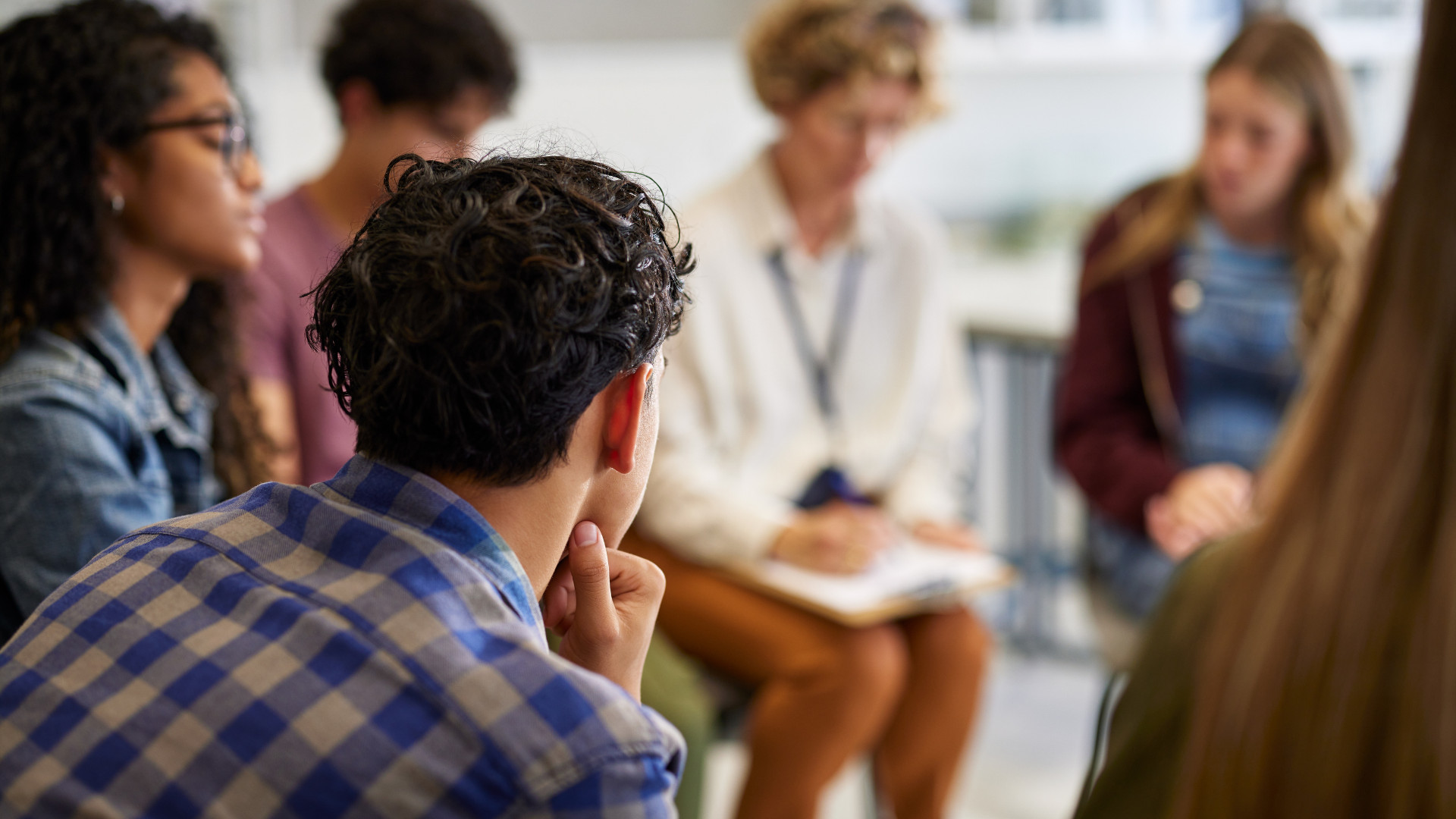 People seated in a circle, listening to a woman with a notepad; group therapy session.