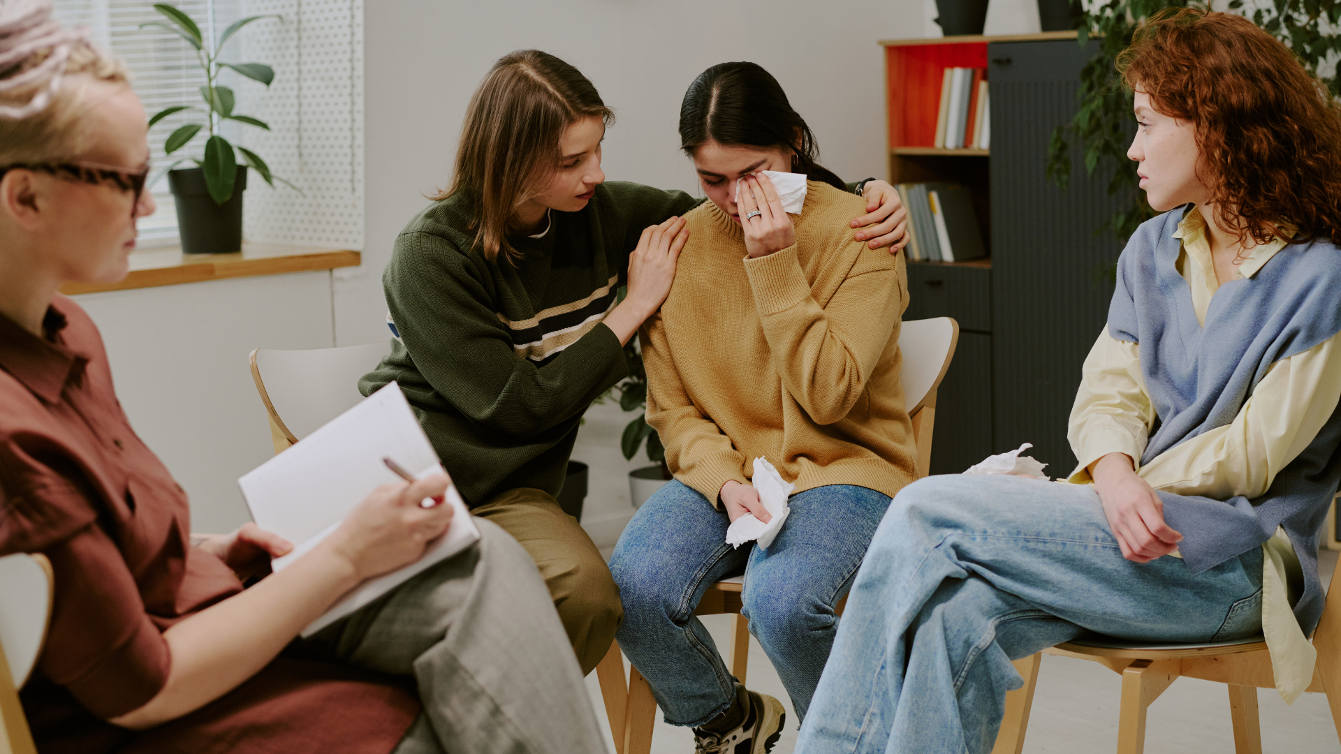 Four people in a circle: one is crying, comforted by another. A person takes notes; another looks on.
