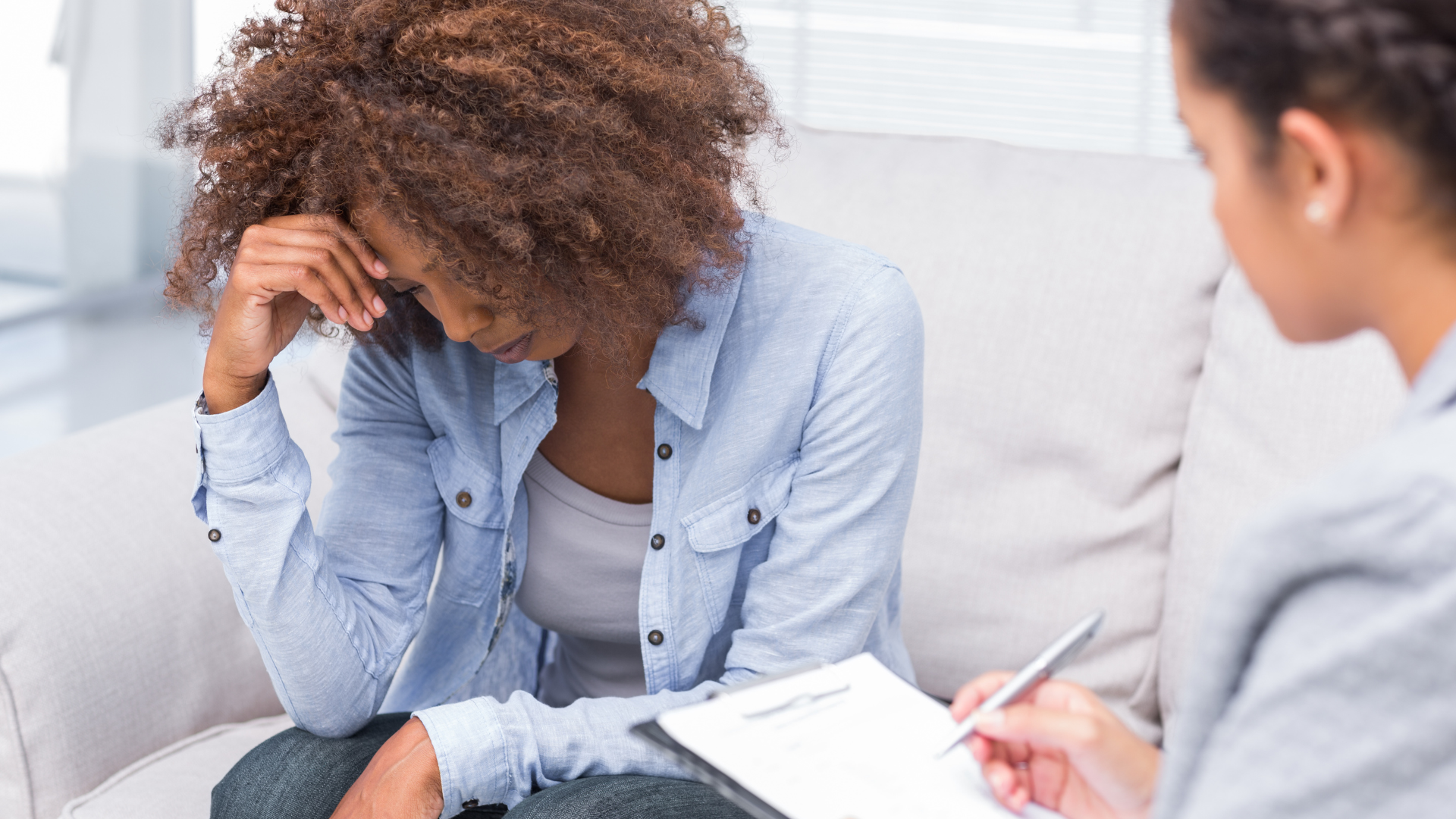 Woman with curly hair looking down, holding her head, talking with a person with a clipboard. Indoors.
