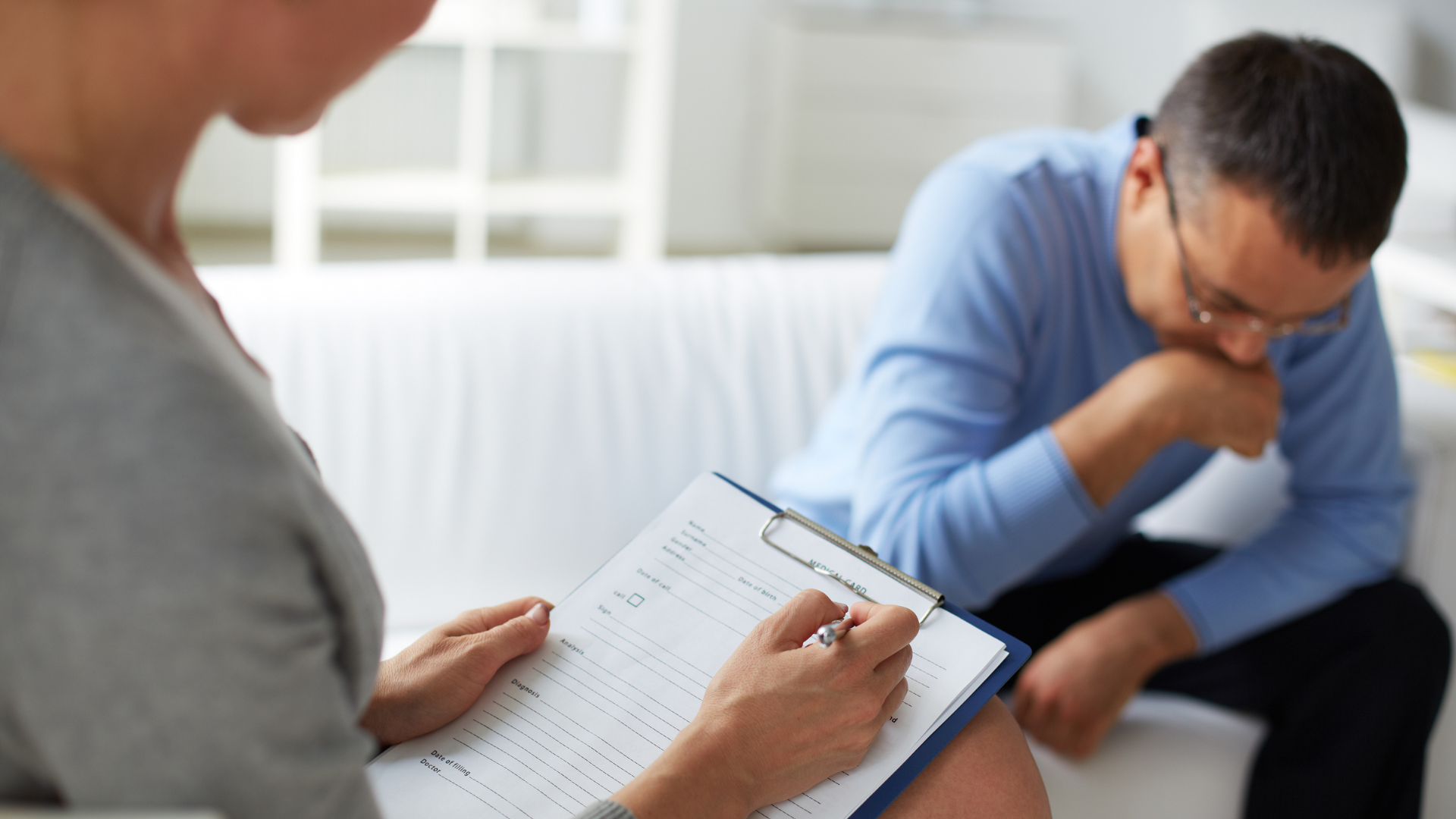 Therapist taking notes while a person in blue shirt sits, looking down, on a white couch.