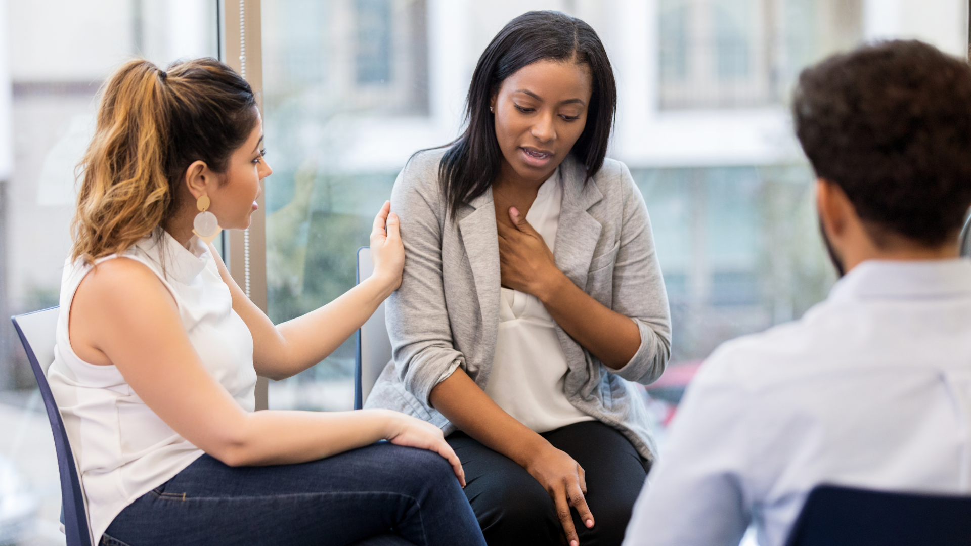 Three people in a counseling session. One person is comforted by another as they hold their chest; a third looks on.