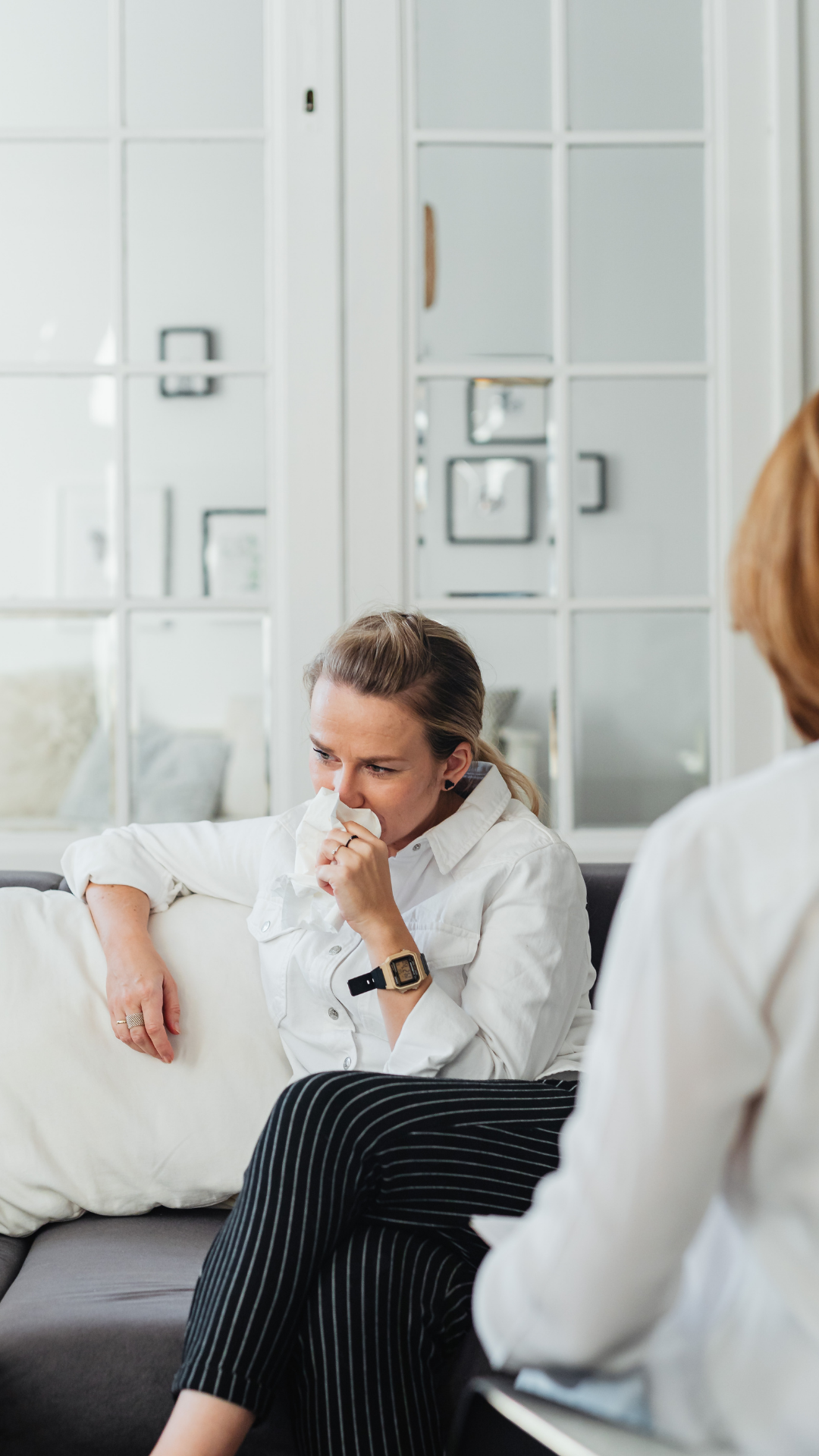Woman on a couch wiping her eyes, talking with a blurred therapist in a light-filled office.