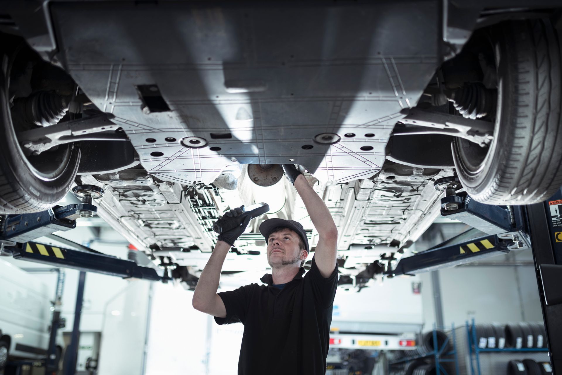 Mechanic working under a car on a lift, wearing gloves and a hat, in a garage.