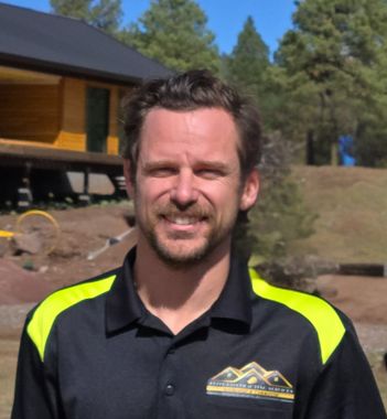 Man smiling, wearing a black shirt with neon yellow shoulder accents; a mountain backdrop.