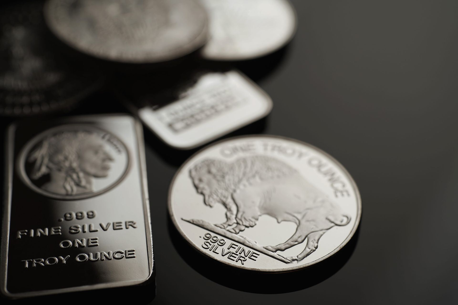 Silver bullion bars and coins on a dark surface, with a buffalo design on one coin