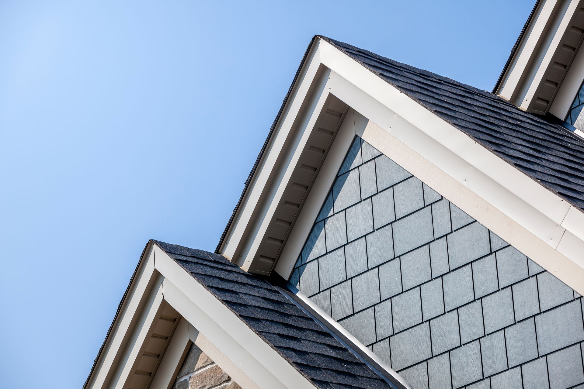 Close-up of a house roof with blue shingles and grey siding against a clear blue sky.