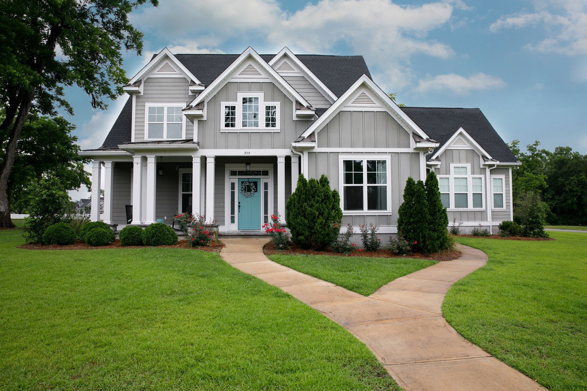 Gray two-story house with blue door, white trim, and a split walkway through green lawn under a cloudy sky.