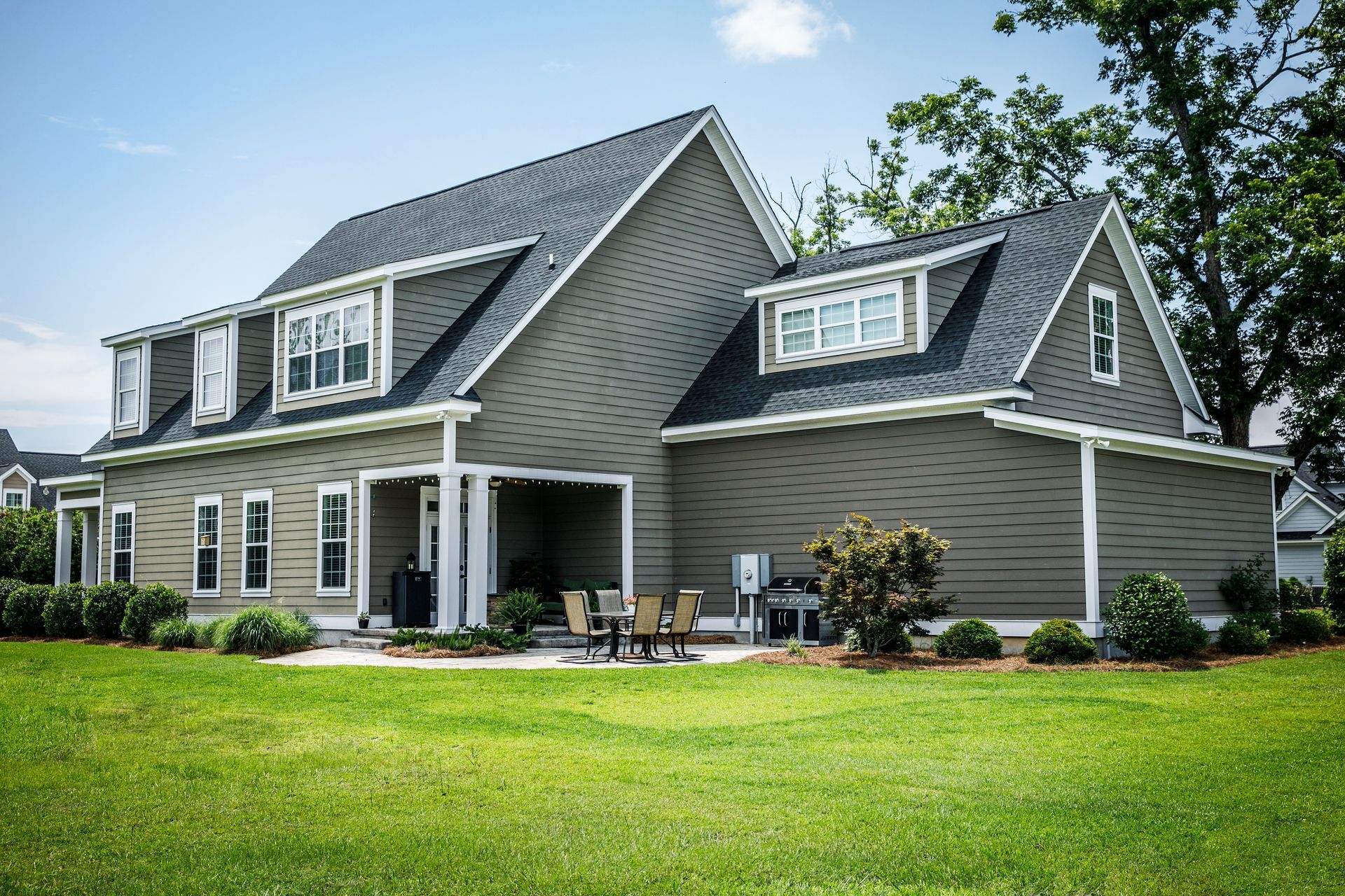 Gray house with a dark roof and white trim, on a green lawn. Patio area with furniture and grill.