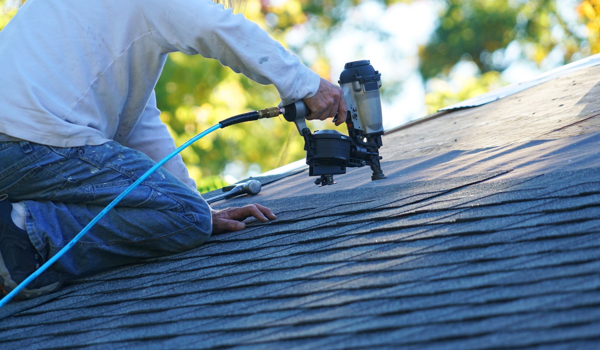Worker kneels on roof installing shingles with pneumatic nail gun.