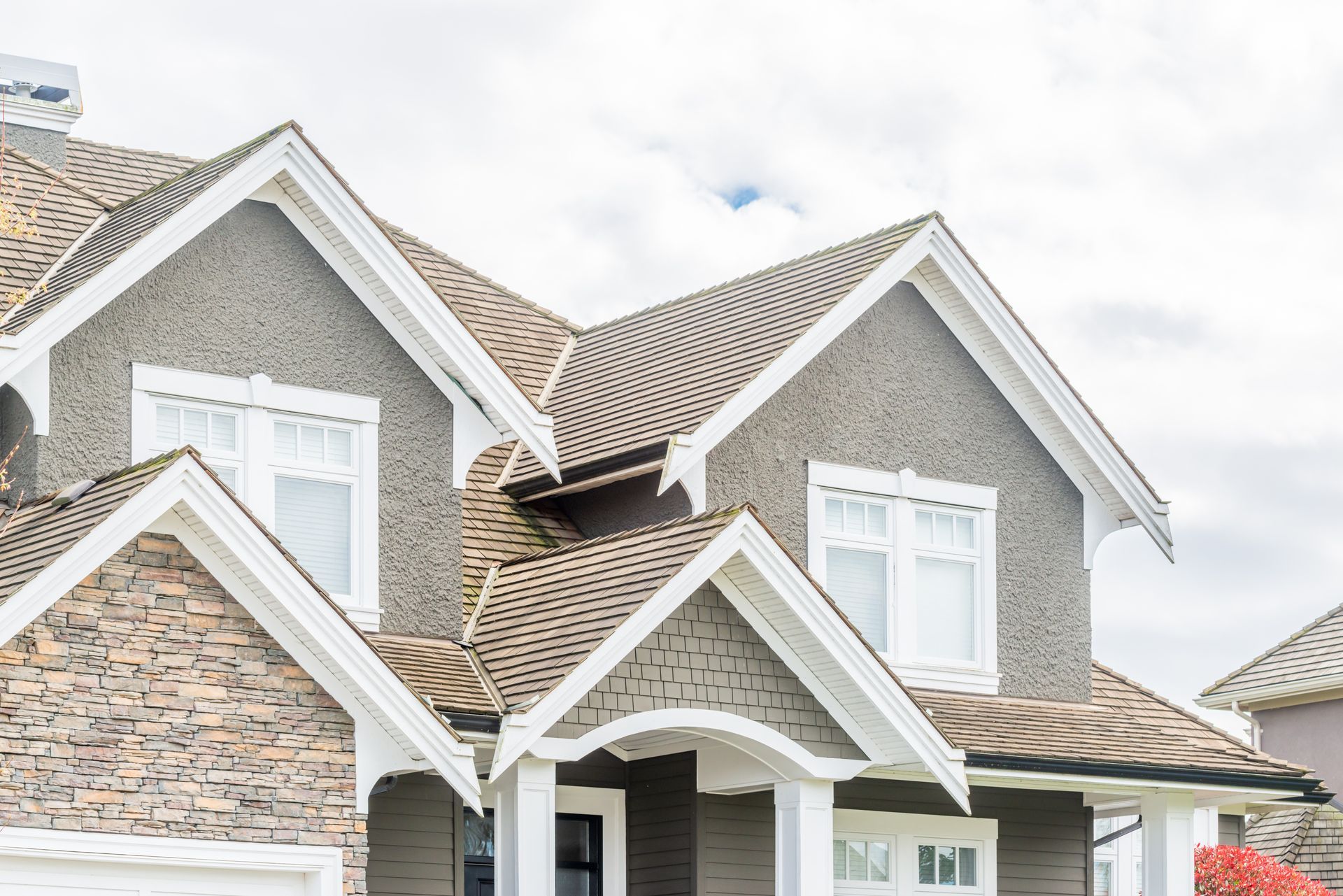 Multi-gabled house with gray siding, stone accents, white trim, and a brown shingle roof against a cloudy sky.