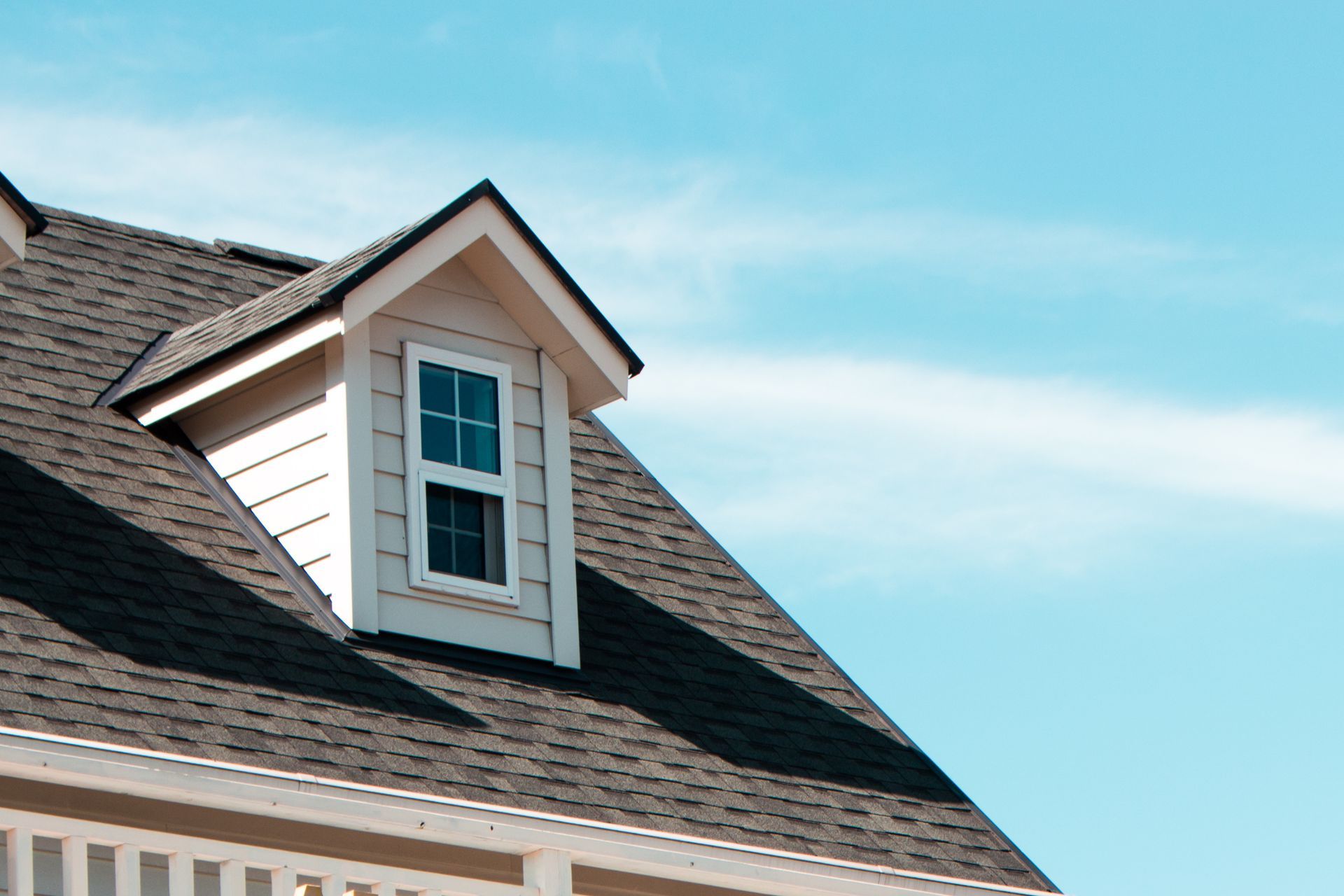 Beige dormer with window on a dark shingle roof against a clear blue sky.