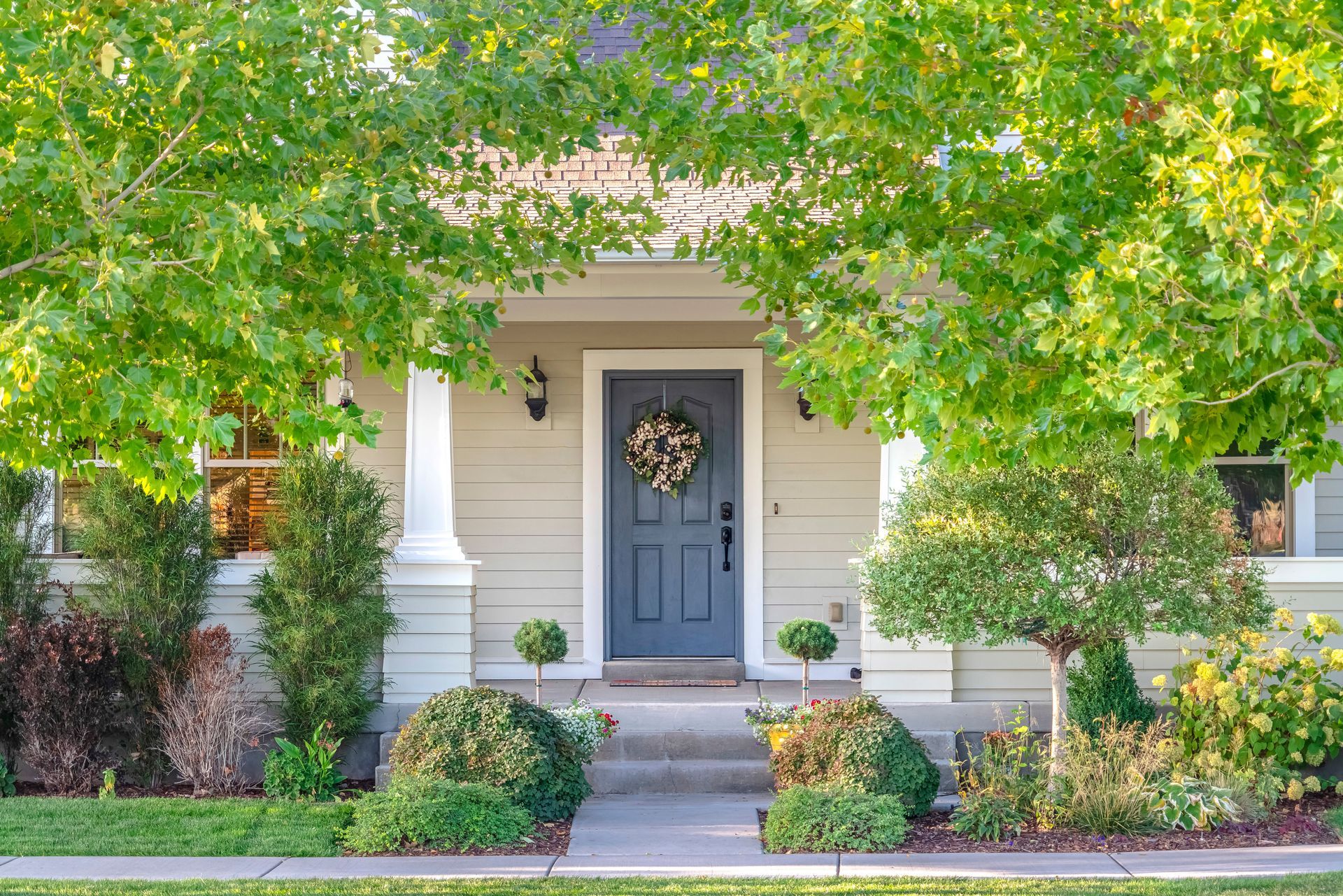Beige house with blue door, wreath, and green foliage.