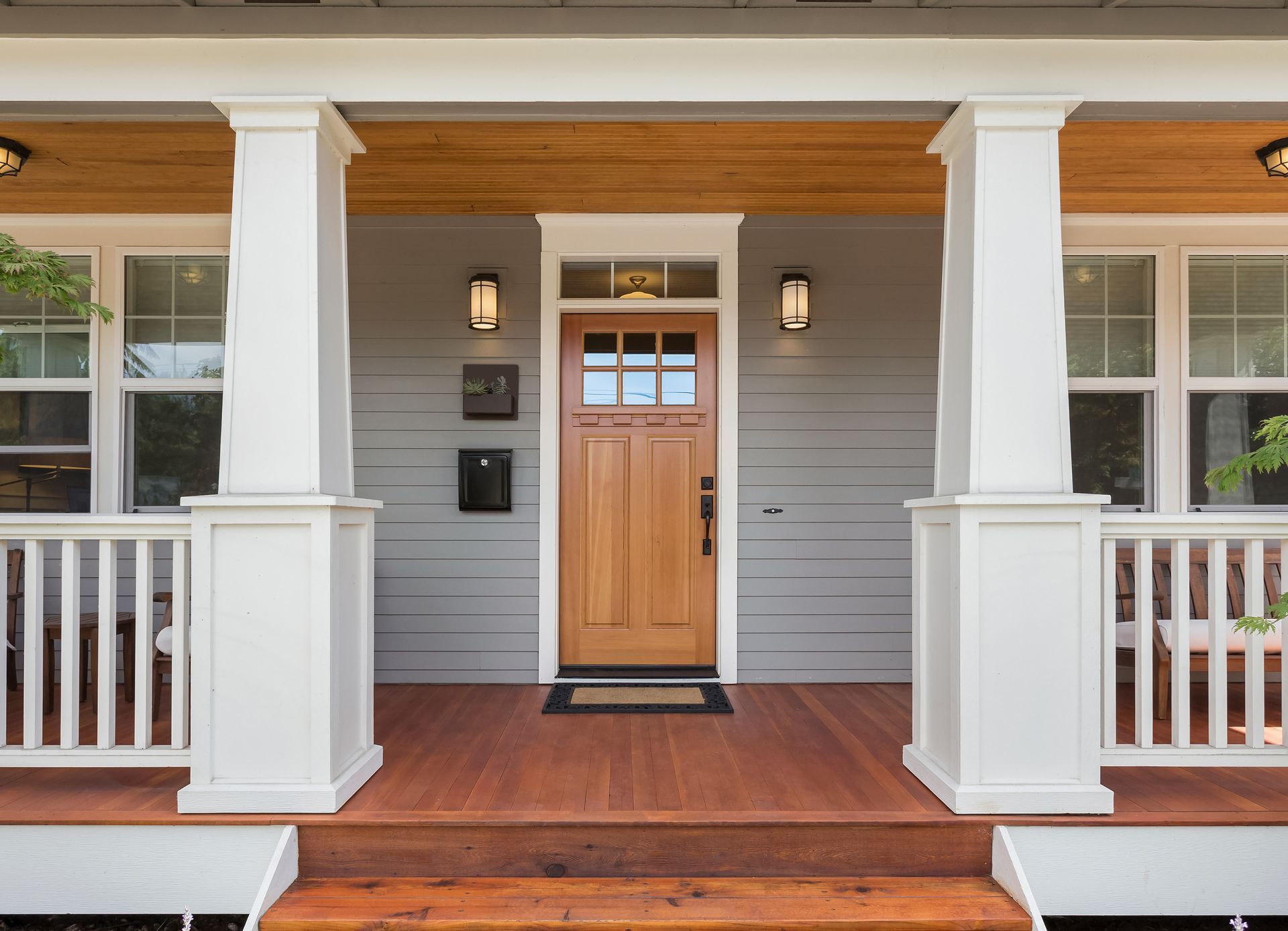 Front porch of a house with wooden door and white pillars. Reddish-brown floor and gray siding.