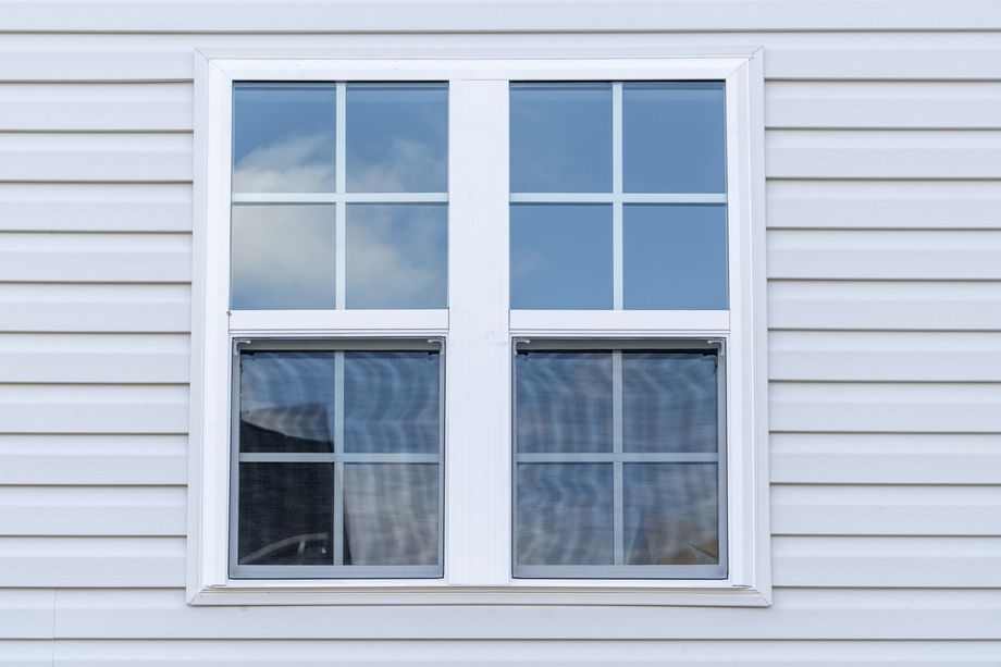 White double-hung window with a grid pattern, set in white siding, reflecting a cloudy sky.