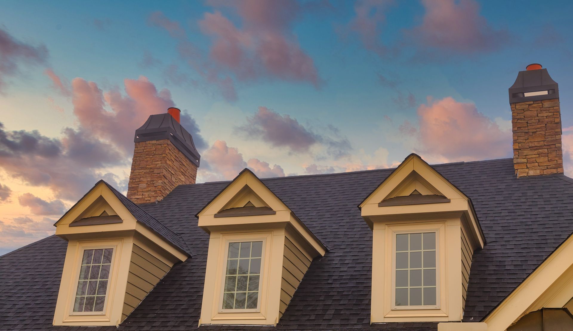A house roof with three dormer windows, two chimneys, and a colorful sunset sky.