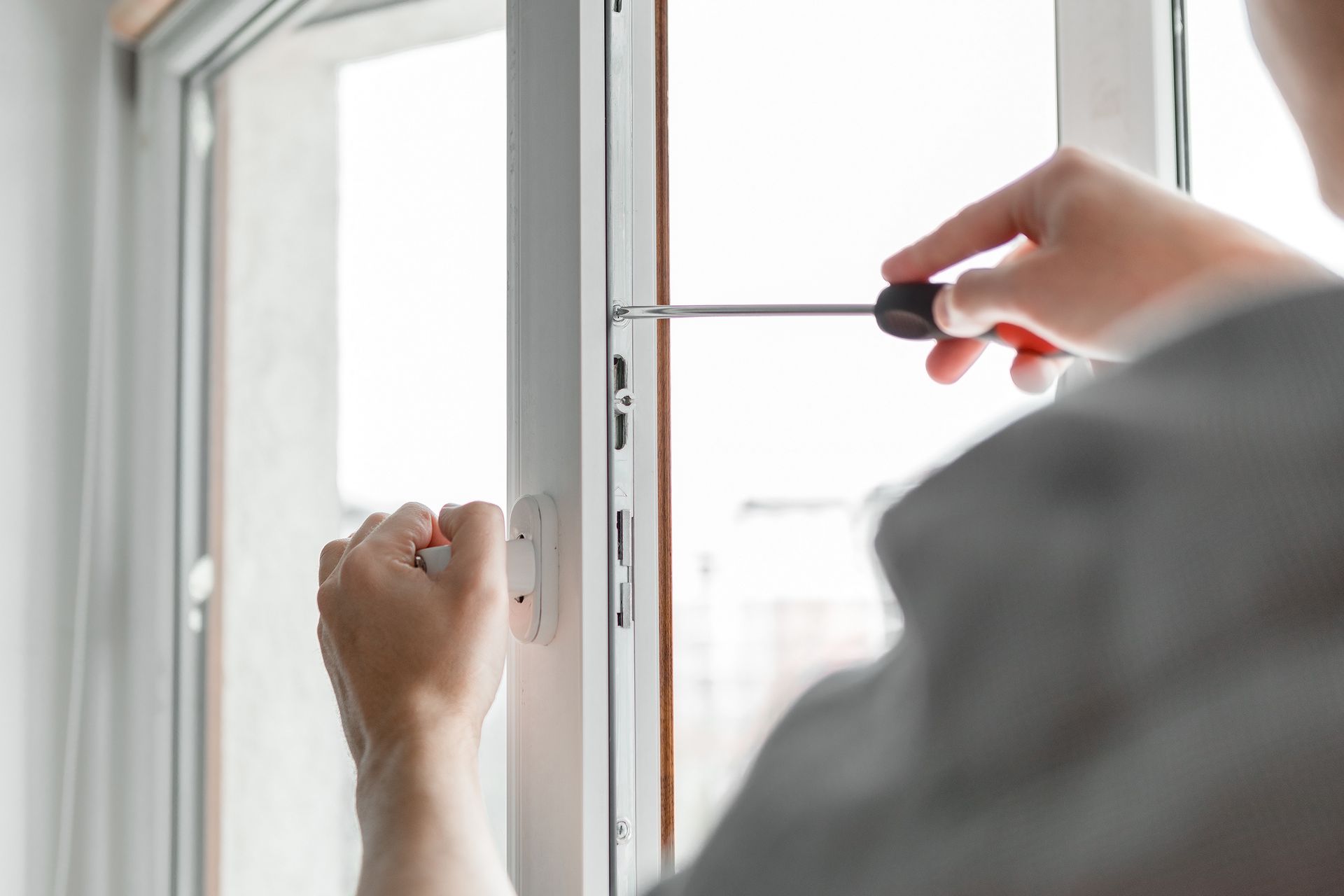 Person using a screwdriver to adjust a white window frame's locking mechanism.