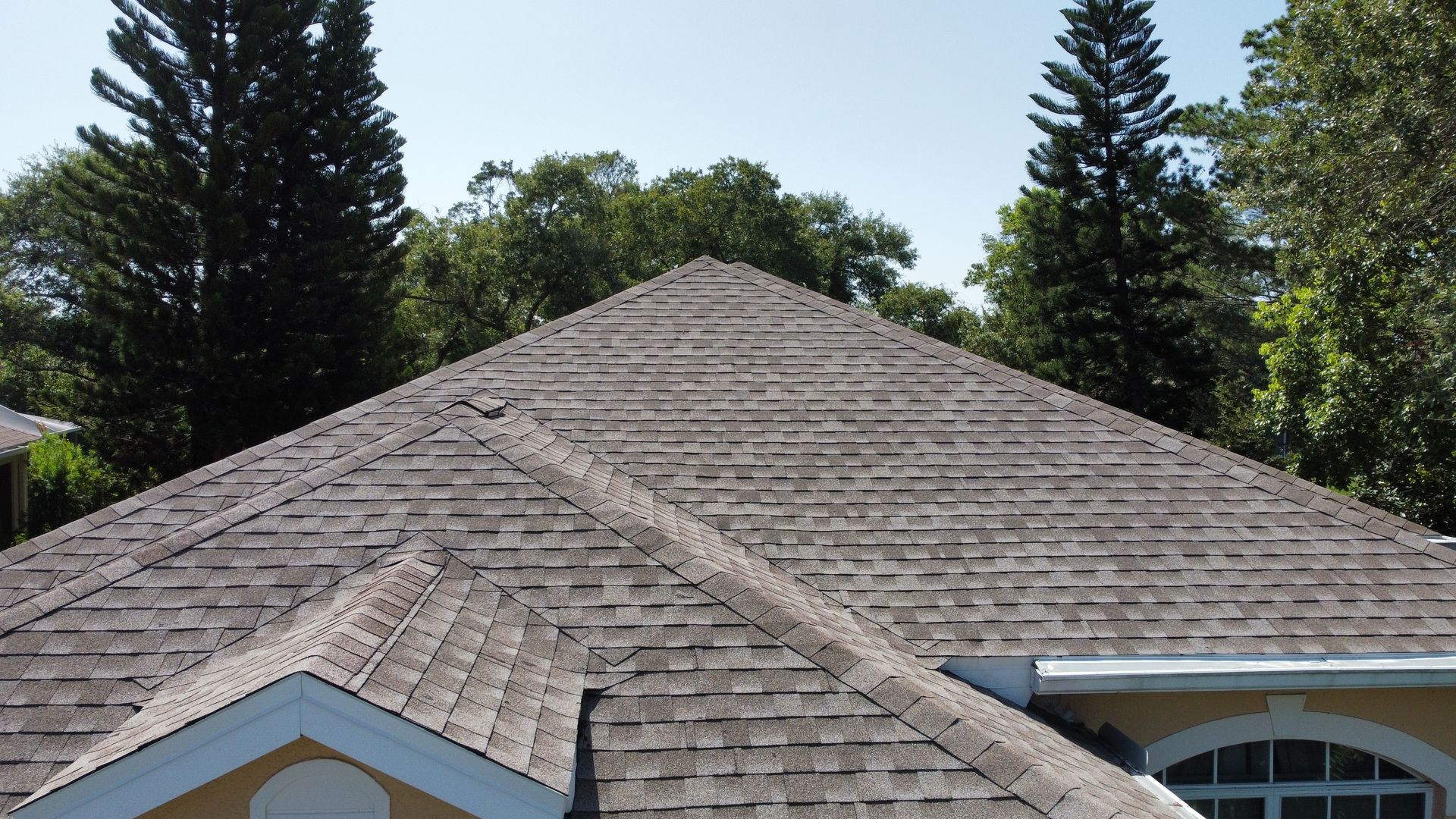 Gray shingled roof of a house, triangular shape, surrounded by green trees under a blue sky.