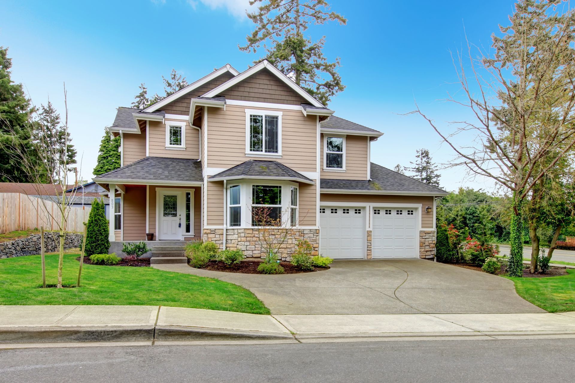 Two-story beige house with white garage doors and a paved driveway under a blue sky.