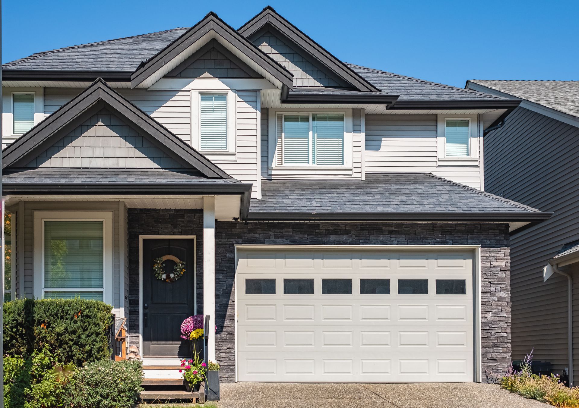 Two-story house with gray siding, dark roof, front door with wreath, and white garage door.
