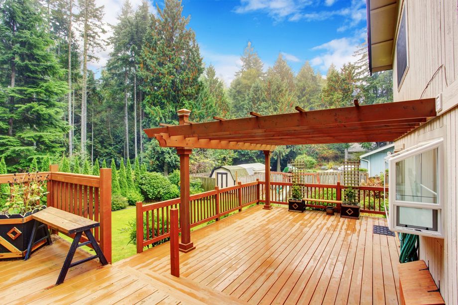 Wooden deck with pergola, overlooking a green lawn and trees, with a building in the background.