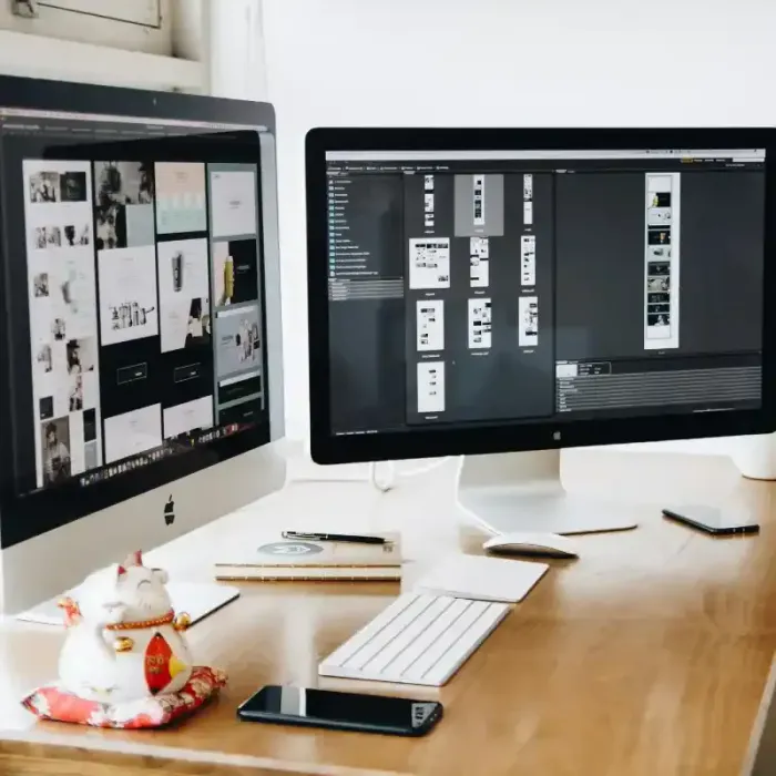 A desk containing two Apple monitors with various photos showcasing the website build process. There is also a white Apple mouse, white Apple keyboard and black cell phone on the desk. There is also a black pen, a book and a decorative porcelain cat.