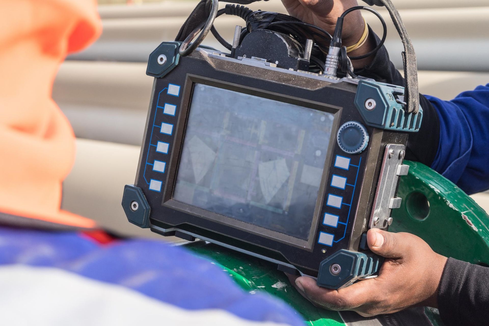 A technician holds a portable industrial testing device displaying data while working on a pipe in an outdoor setting.