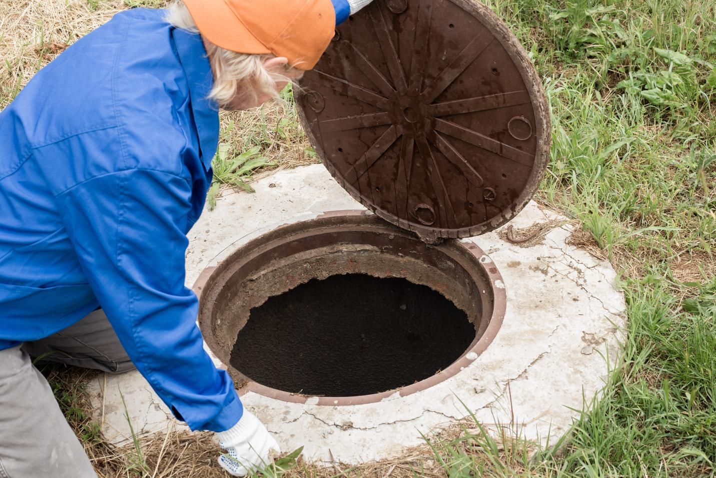A person in a blue jacket and orange hat kneels on grass, lifting the heavy metal cover of a dark underground septic tank.