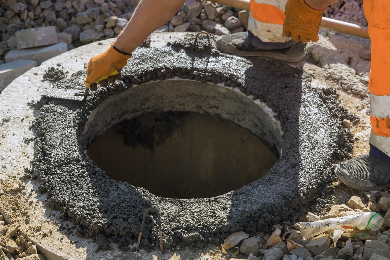 A construction worker in high-visibility gear uses a trowel to apply wet cement around a circular manhole opening.