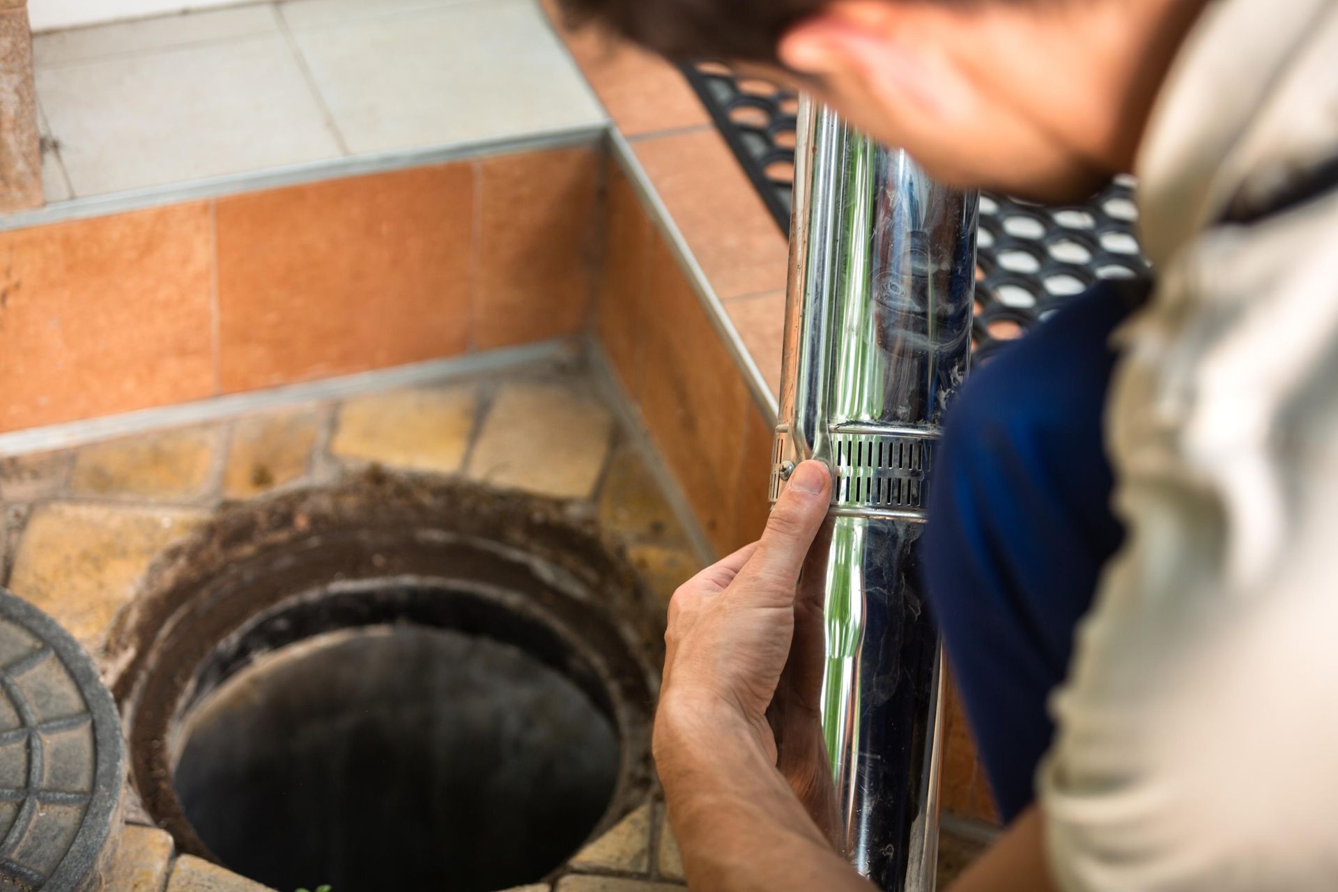 A person works on a shiny metal pipe near an open circular drain in a tiled outdoor area.
