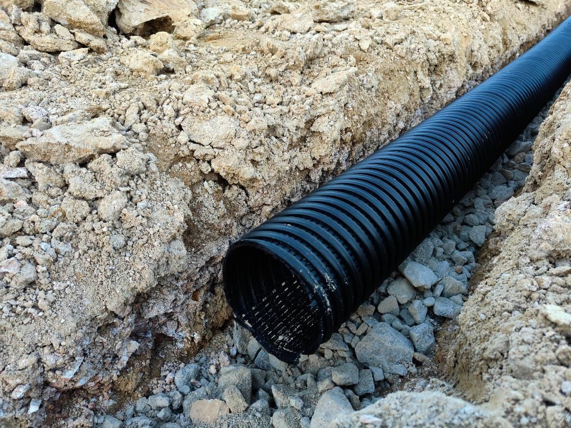 A black corrugated drainage pipe lies in a shallow gravel-filled trench cut into dirt ground.