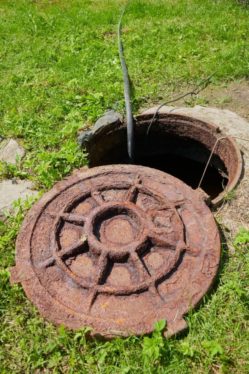 A rusty, circular iron manhole cover lies open on grass, with a dark pipe protruding from the exposed shaft.