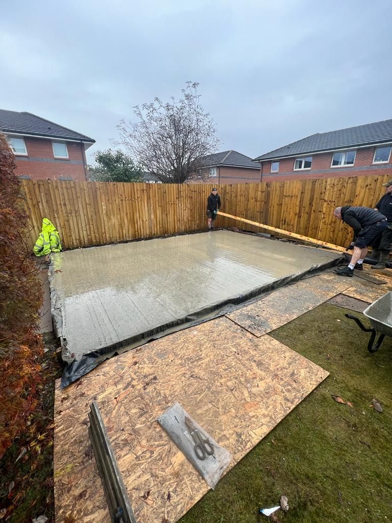 two men are working on a concrete floor in a backyard for a garden room