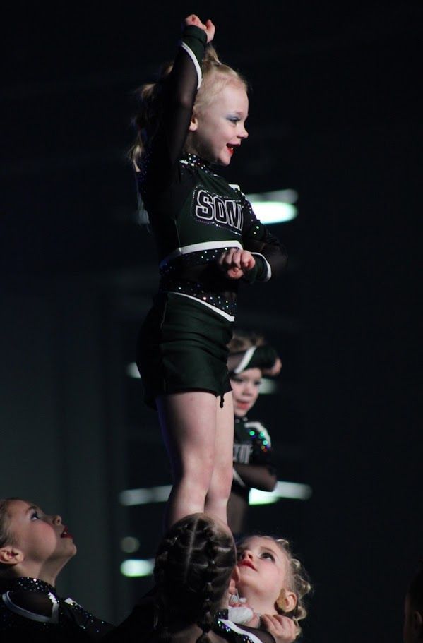 Young cheerleader atop teammates in dark green uniform, arms raised.