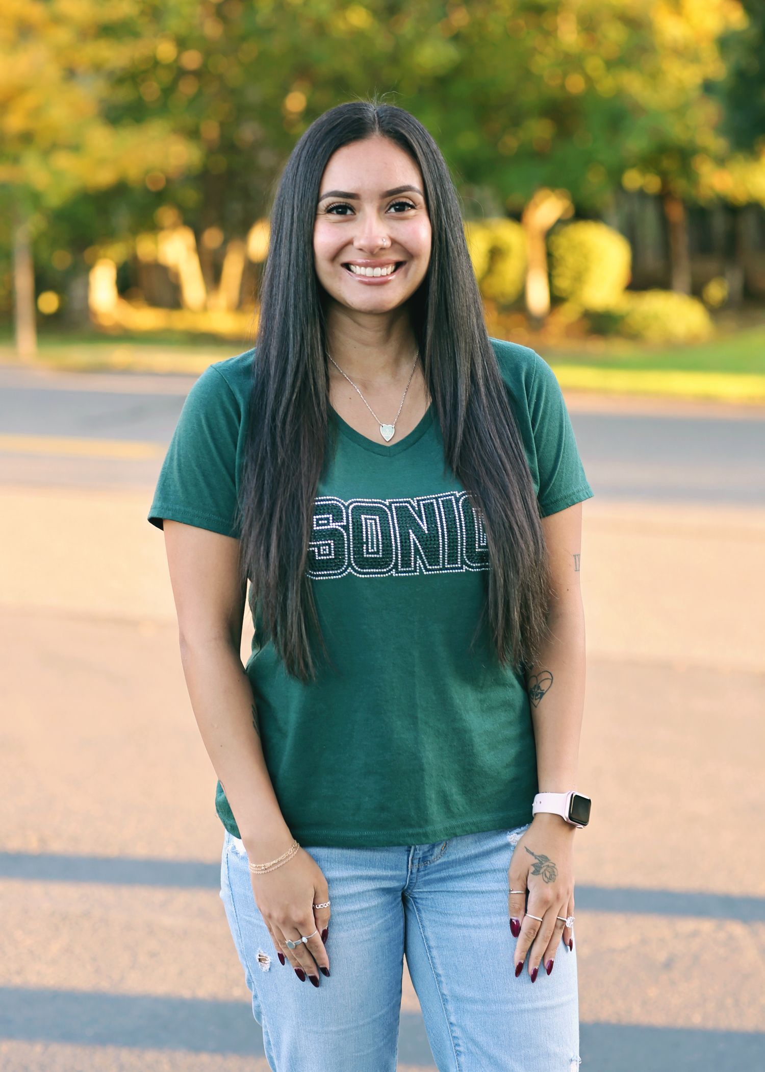 Woman with long dark hair, wearing green shirt and jeans, smiling outdoors.