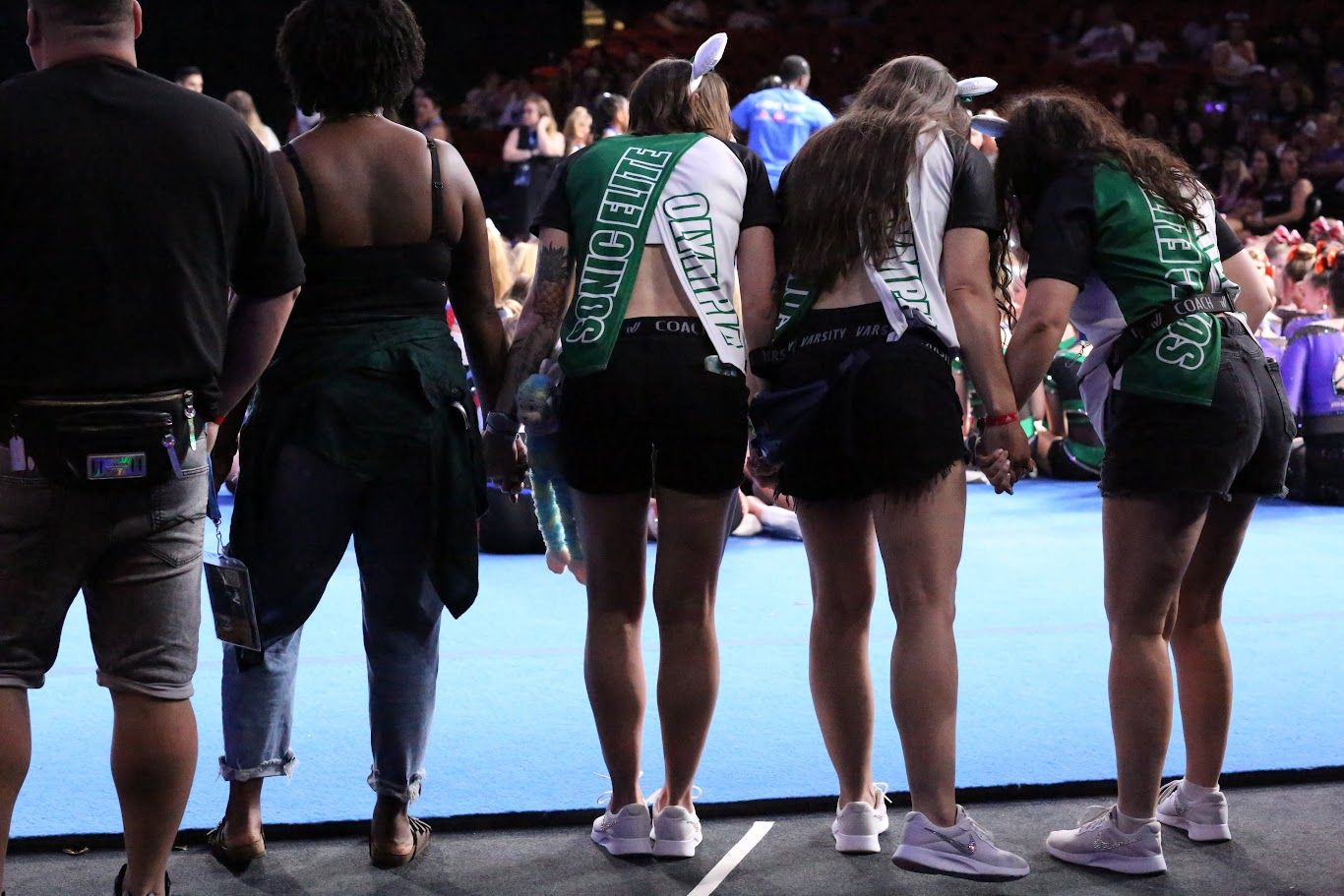 People in a crowd wearing green and white cheerleader outfits, holding hands.