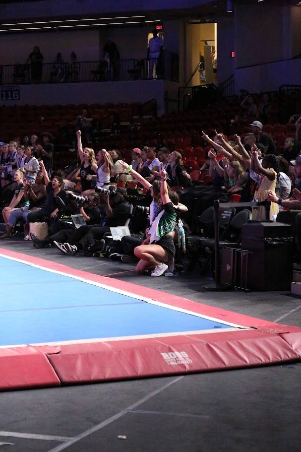 Cheerleaders cheering, arms raised, in arena. Others watching, many with hands raised.