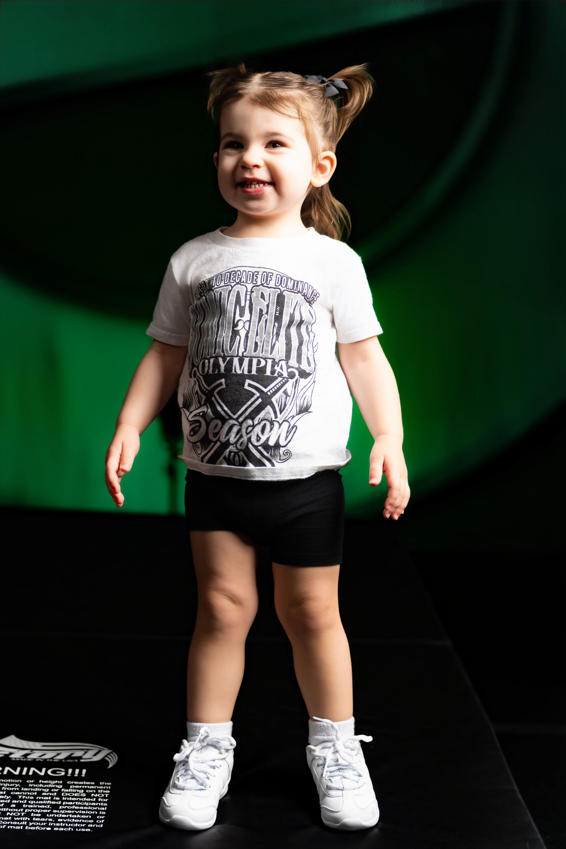 A young cheerleader kneels, arms crossed, smiling. Wearing green and black uniform on a stage.