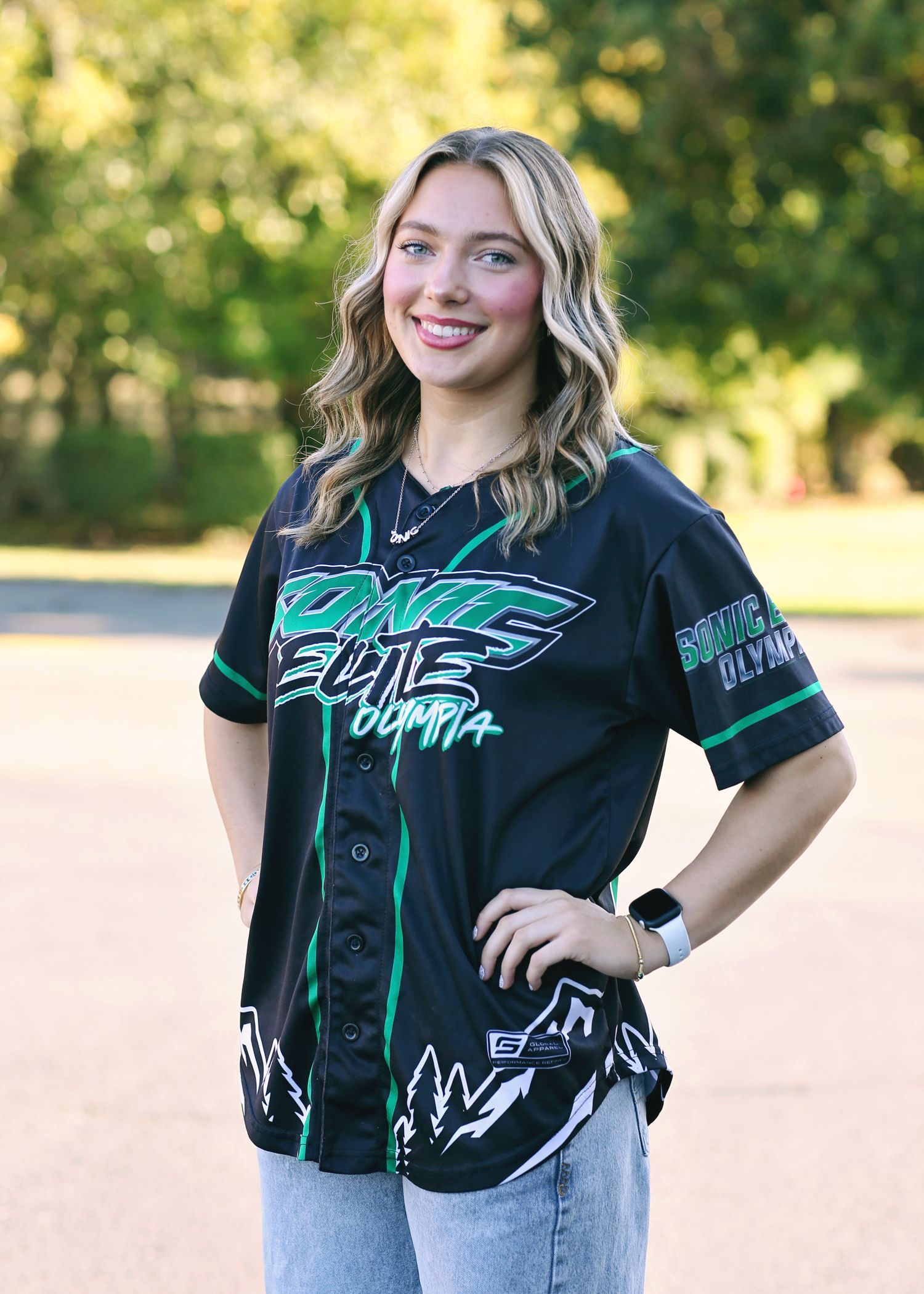 Woman wearing a black and green baseball jersey, smiling, hands on hips, standing outdoors.