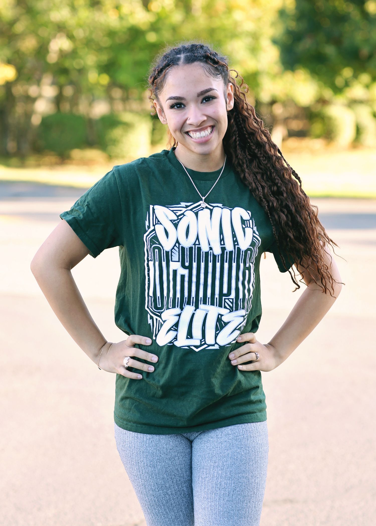 Young woman in green shirt and leggings smiling, hands on hips, outdoor setting.
