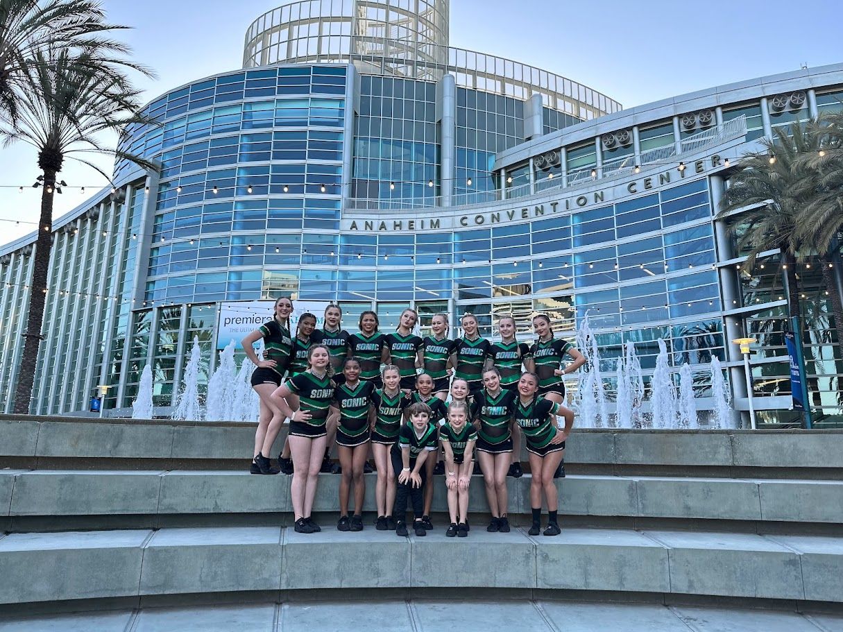 Cheerleading team poses in front of a modern building with a fountain; they wear green and black.