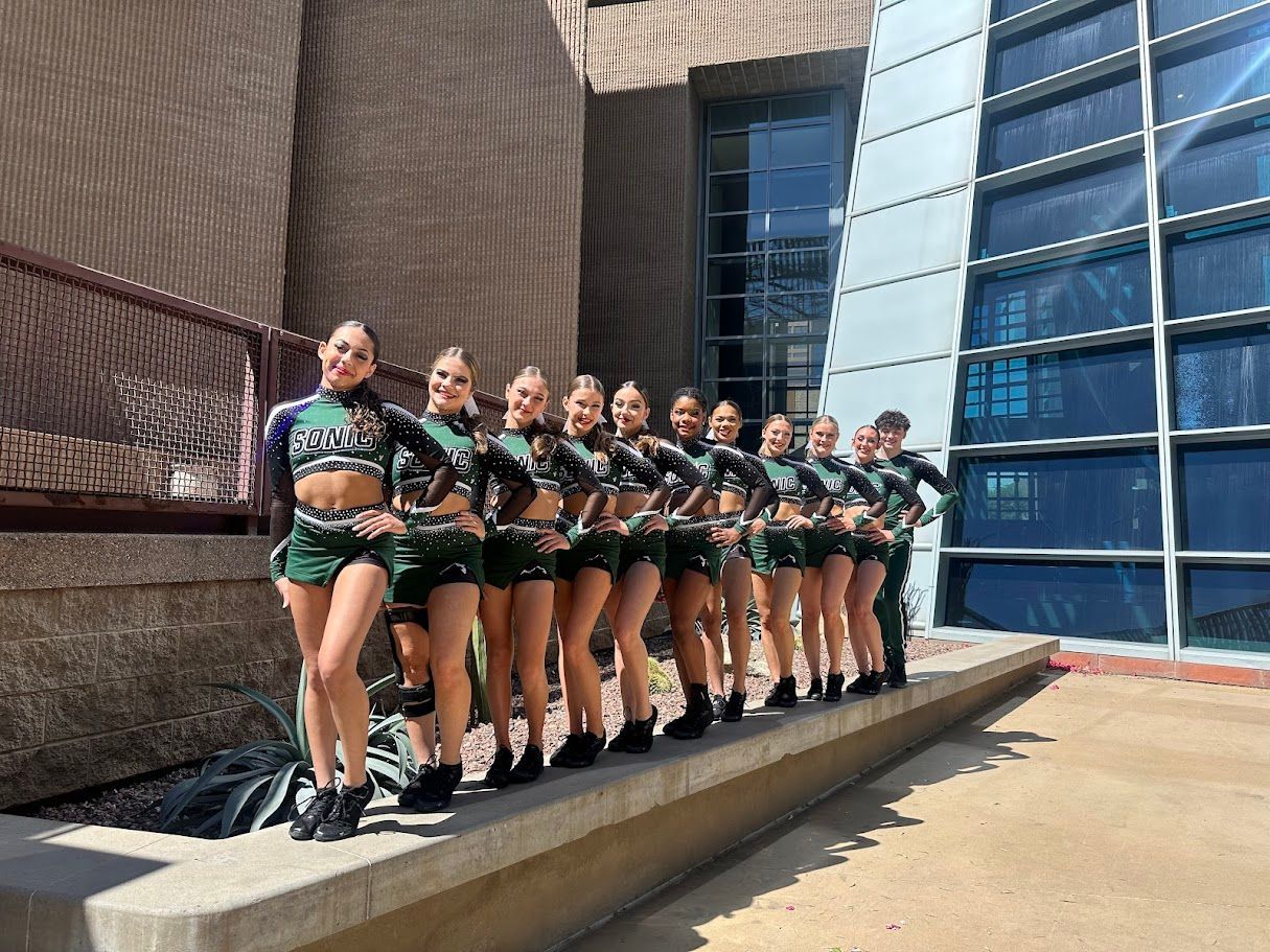 A cheerleading team in green and black uniforms poses outside a building.