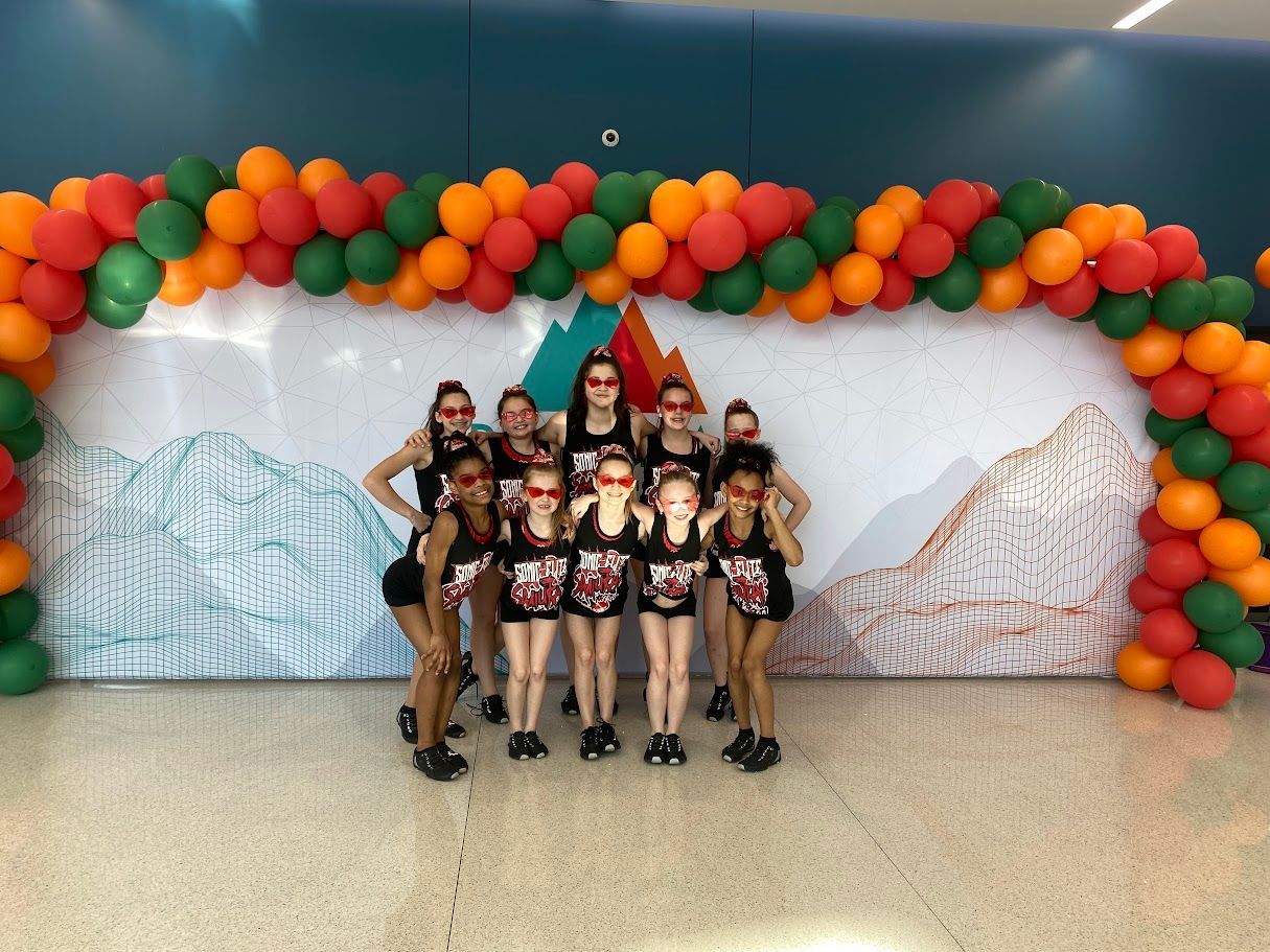 A dance team poses in front of a mountain-themed backdrop with a balloon arch.