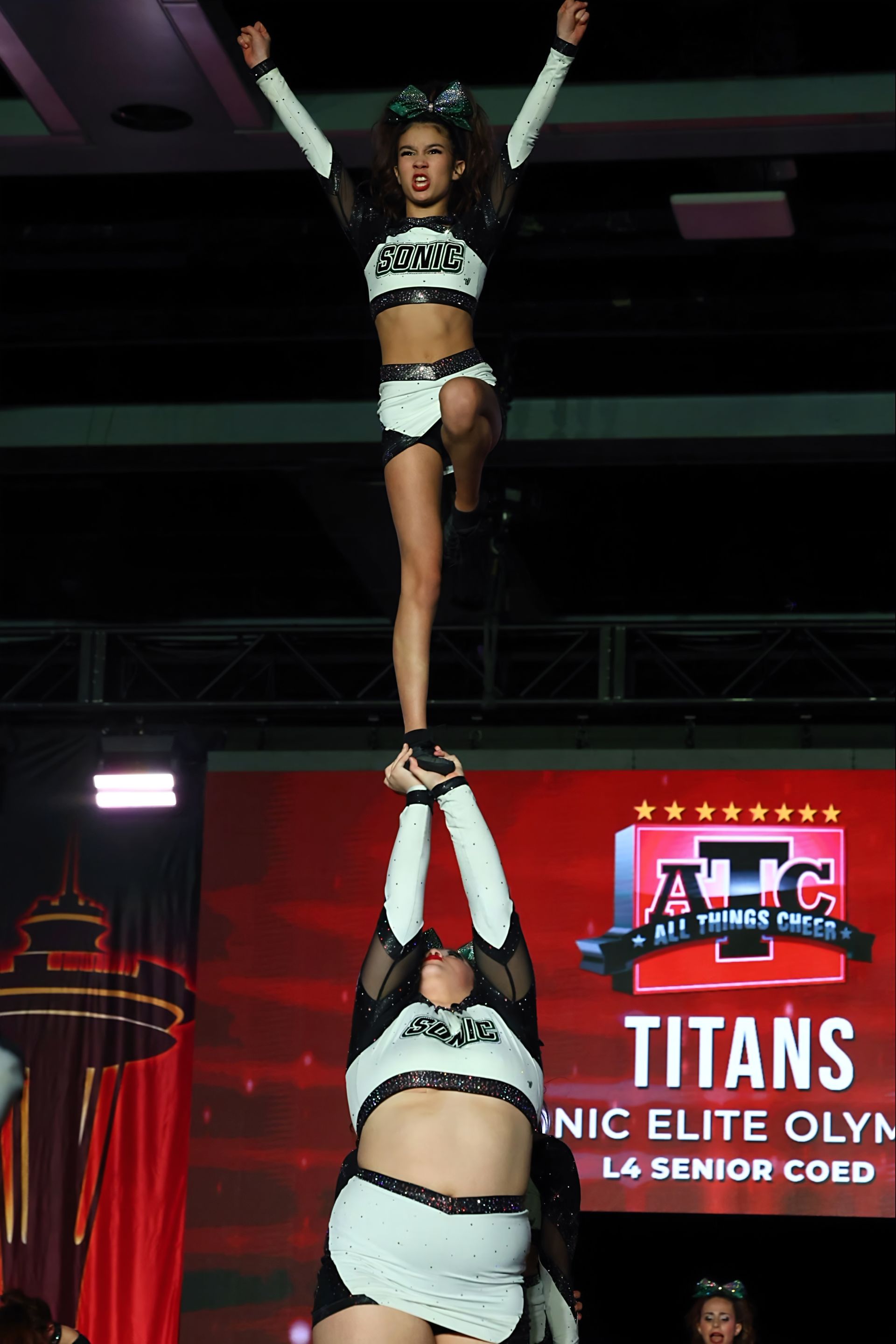 Cheerleaders performing a stunt. A flyer does a split high in the air. Another flyer does a bent leg lift.