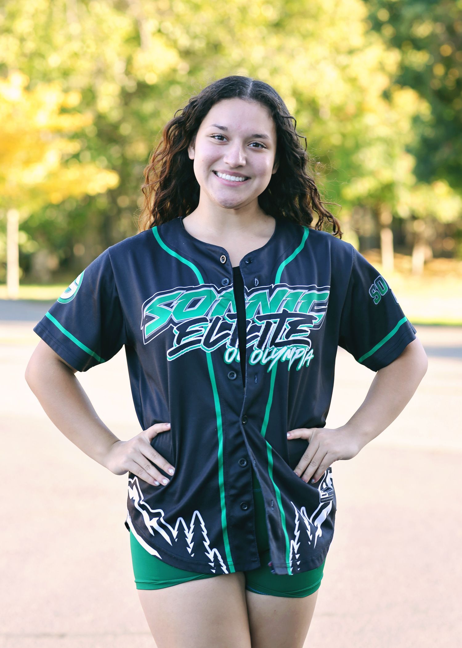 Woman with curly hair smiling, wearing a black and green jersey, hands on hips.