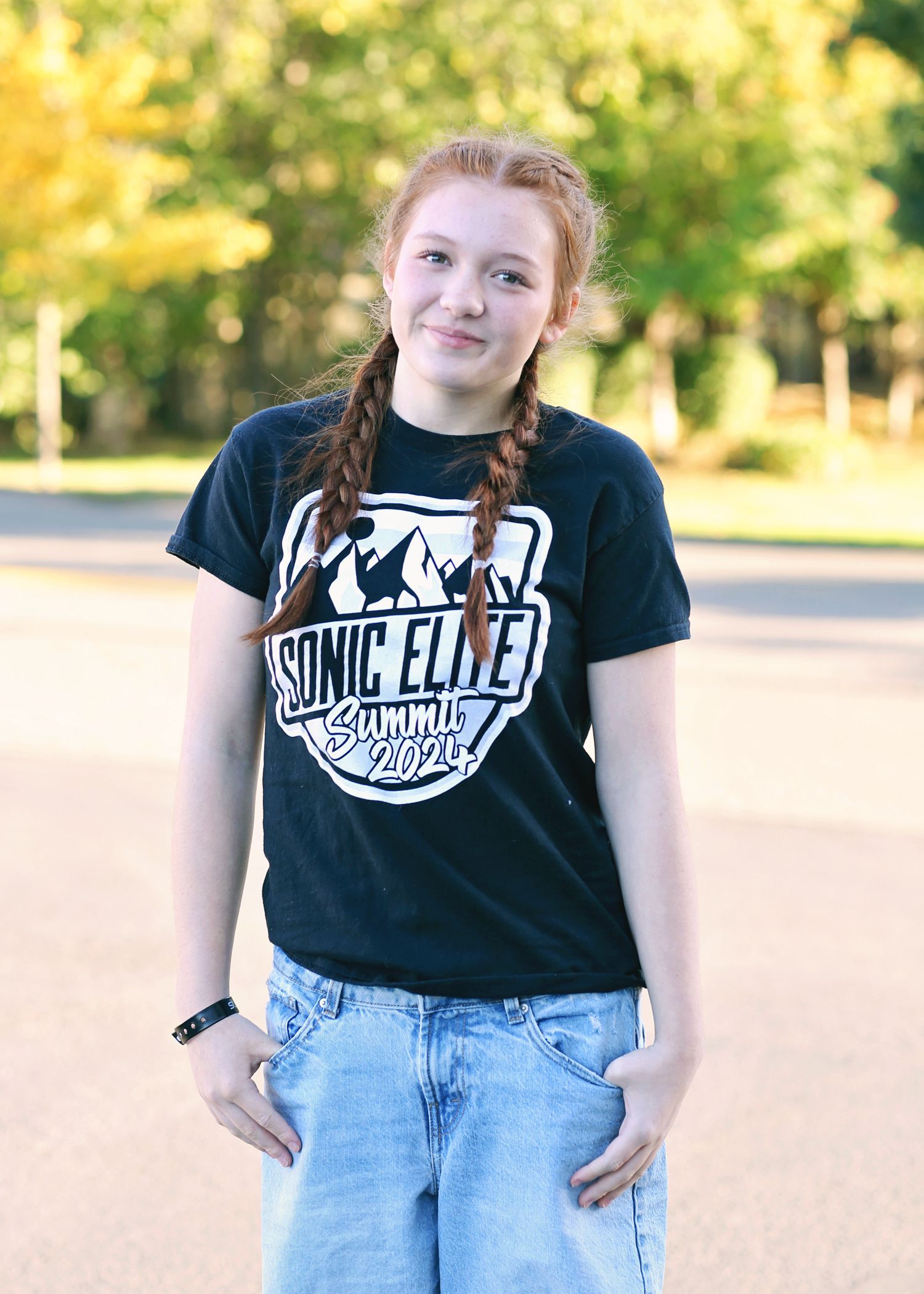 Teen girl with red braids, wearing a black t-shirt, blue jean shorts, smiles. Outdoors.