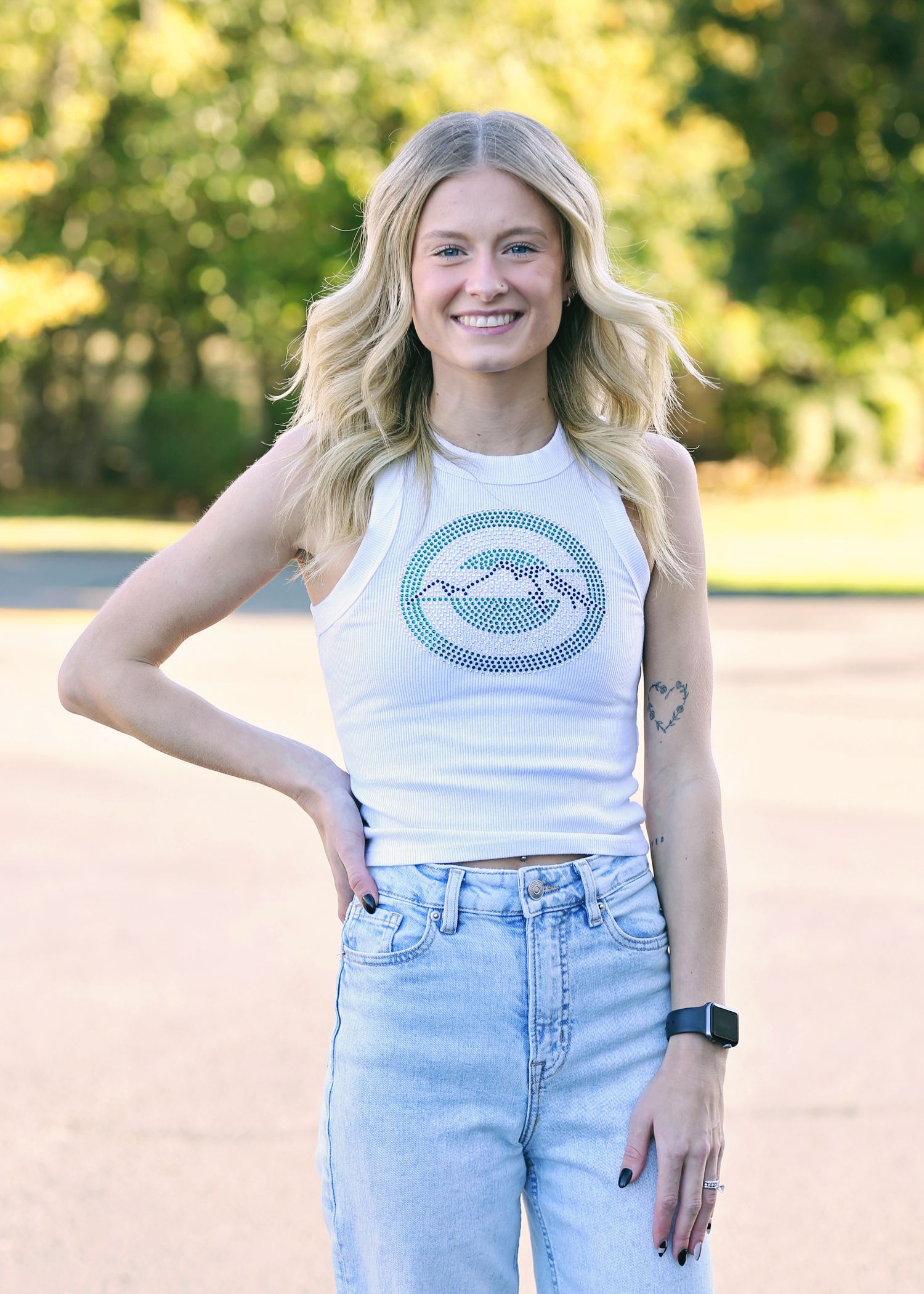 Woman with blonde hair wearing a white tank top and light wash jeans smiles, with one hand on her hip.