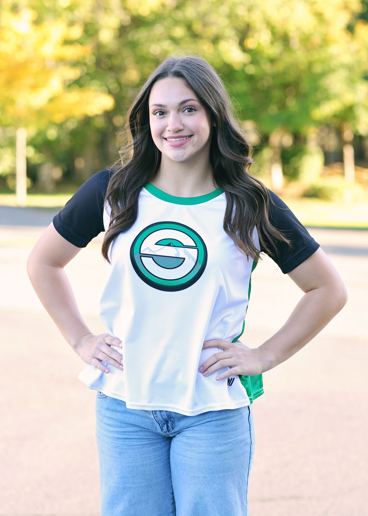 Woman in white shirt with green logo, jeans, hands on hips, smiling, outside.