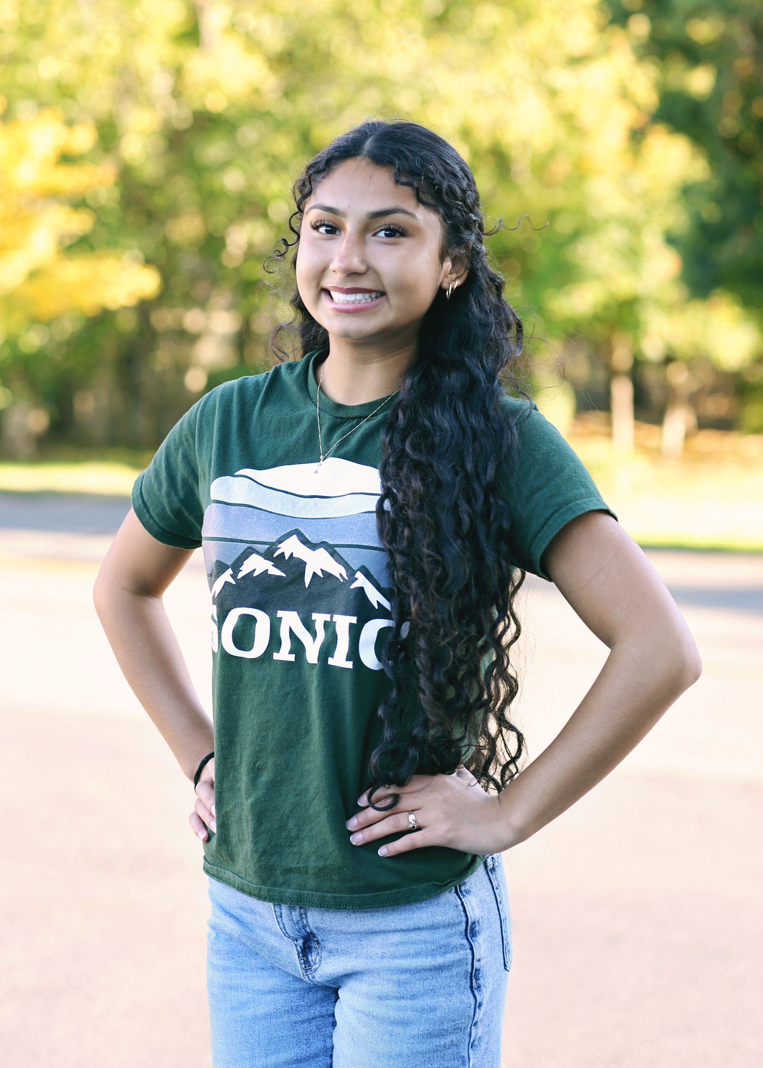 Young woman with long dark curly hair, wearing a green t-shirt and blue jeans, smiling with hands on hips. Outdoors.