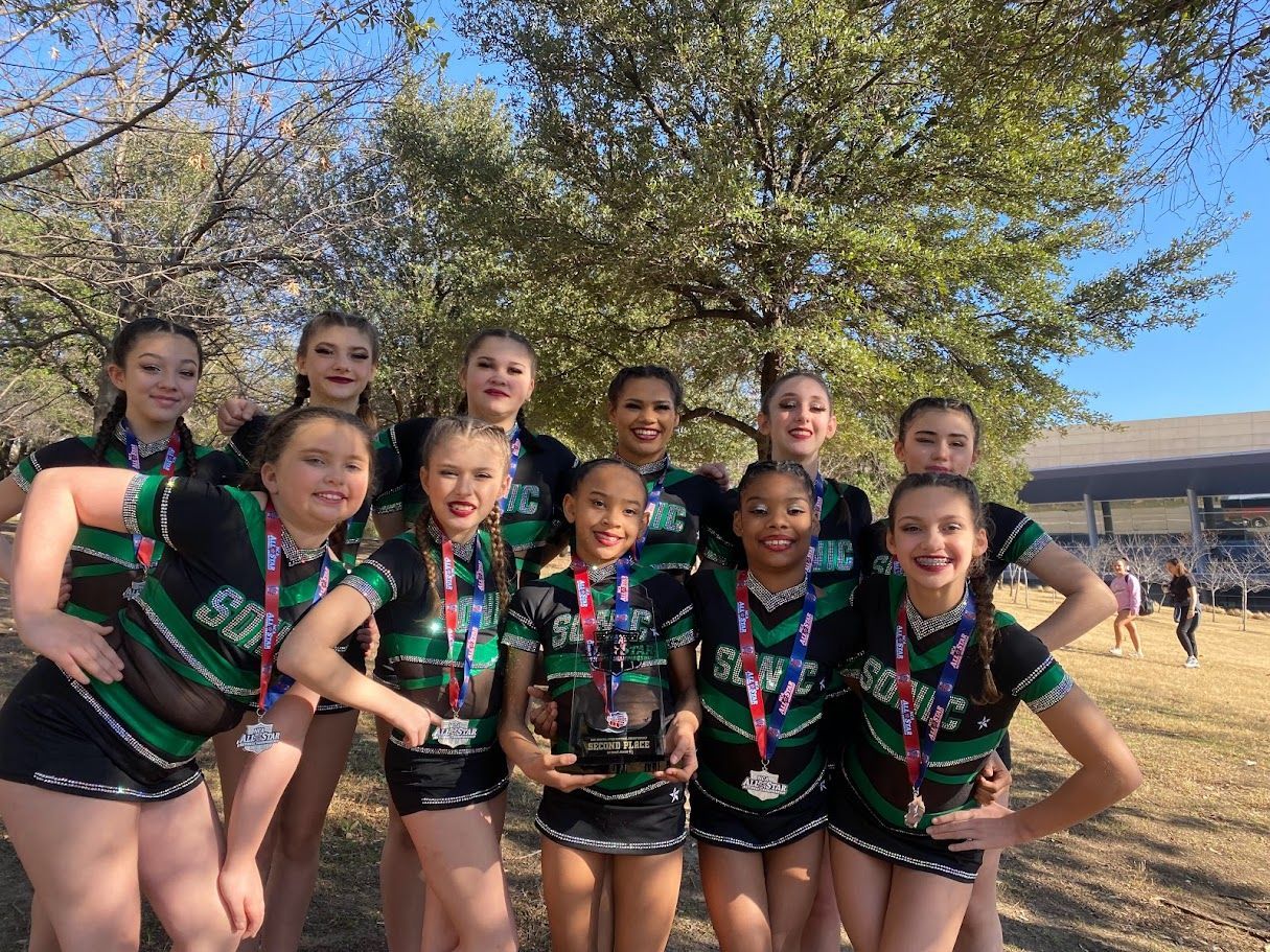 Cheerleaders in black and green uniforms pose outdoors with trophies and medals.