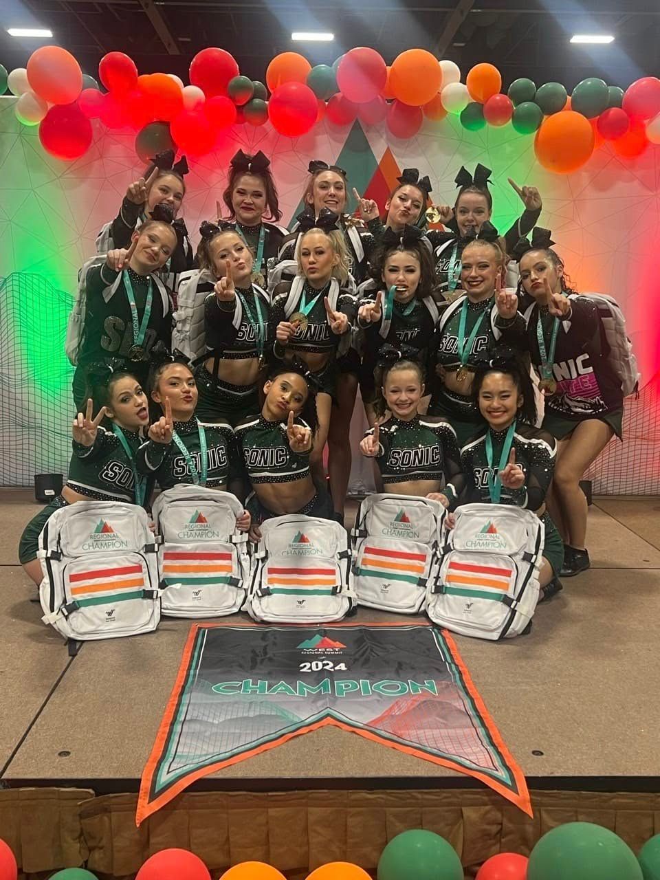 Cheerleading team posing with medals and backpacks, on stage with balloons. Team wears green/black outfits, smiles.